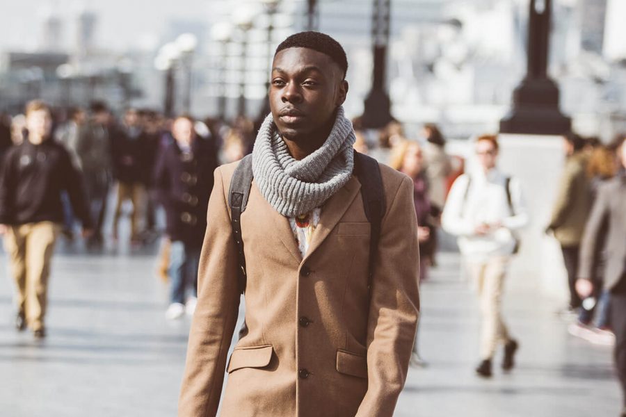 A stylish man wearing a brown coat and scarf walking on a busy city street with people in the background.