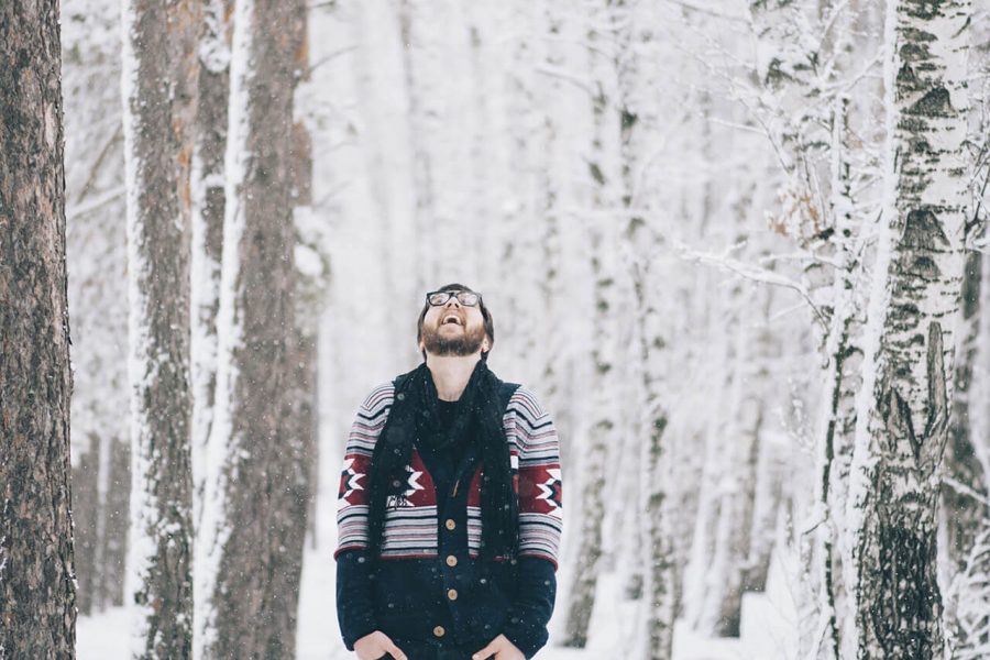 A man wearing a winter jacket stands in a snow-covered forest, looking up with a smile on his face.
