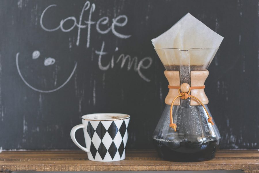 A coffee mug with a black and white diamond pattern next to a glass coffee maker with a wooden collar on a wooden surface.