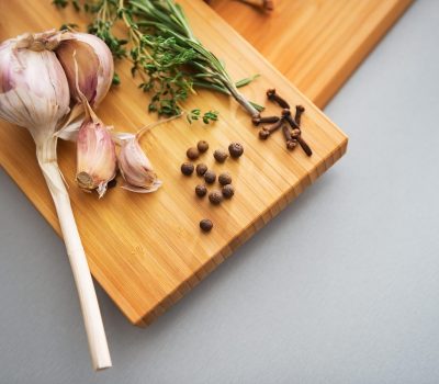 A wooden cutting board with garlic cloves, fresh herbs, cloves, and peppercorns arranged on it.