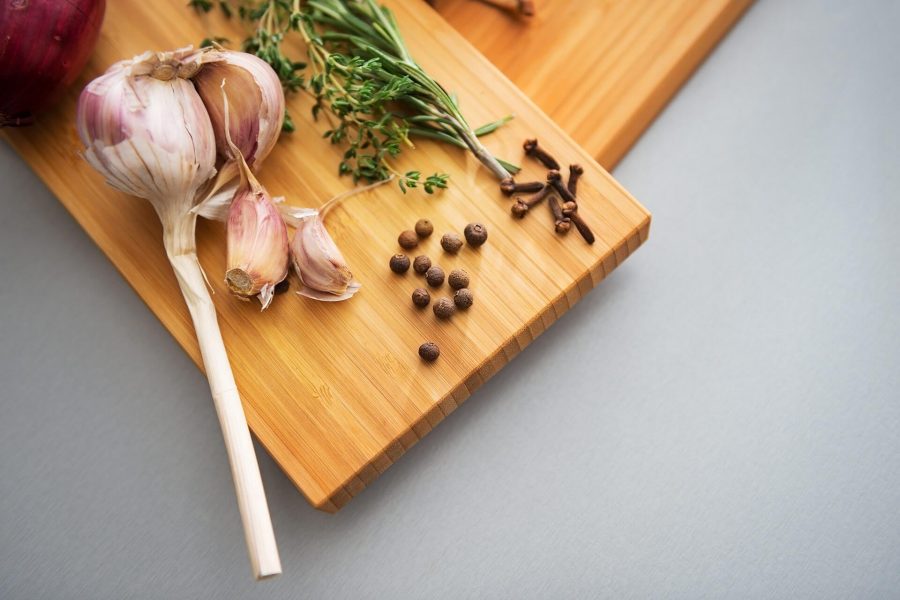 A wooden cutting board with garlic cloves, fresh herbs, cloves, and peppercorns arranged on it.