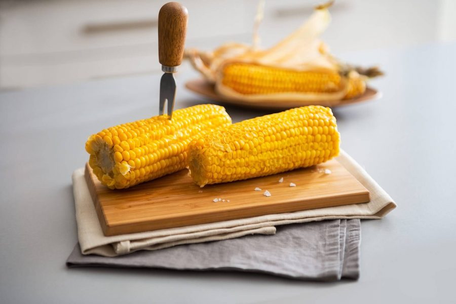 Two grilled corn cobs with wooden handles on a cutting board, with more corn in the background.