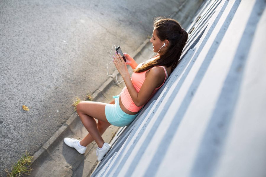 A young woman in casual summer clothes sitting against a wall, focused on her phone outdoors.