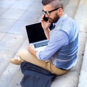 A man with glasses and a beard sitting on outdoor steps, talking on a phone, with a laptop on his lap.