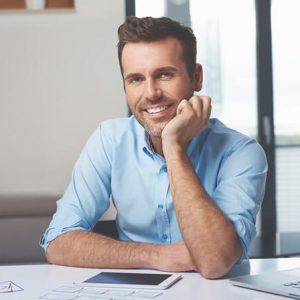 A smiling man in a blue shirt sitting at a desk with documents and a tablet, appearing relaxed and approachable.