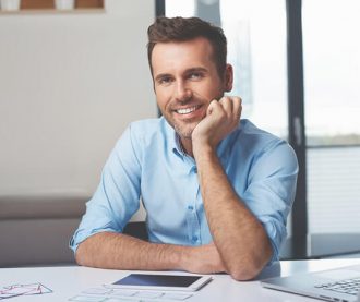 A smiling man in a blue shirt sitting at a desk with documents and a tablet, appearing relaxed and approachable.