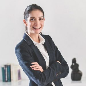 A confident businesswoman in a black blazer and white shirt smiling with arms crossed in a professional setting.