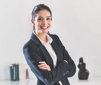 A confident businesswoman in a black blazer and white shirt smiling with arms crossed in a professional setting.