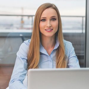A professional woman with long blonde hair, wearing a light blue shirt, sitting and smiling in front of a laptop with a modern office background.