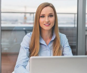 A professional woman with long blonde hair, wearing a light blue shirt, sitting and smiling in front of a laptop with a modern office background.