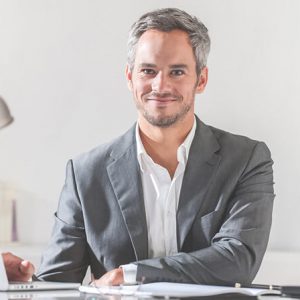 A confident man in a gray suit sitting at a desk with a slight smile.