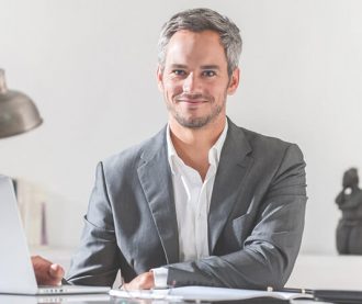 A confident man in a gray suit sitting at a desk with a slight smile.