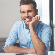 A smiling man in a light blue shirt sitting indoors with his hand resting on his chin.