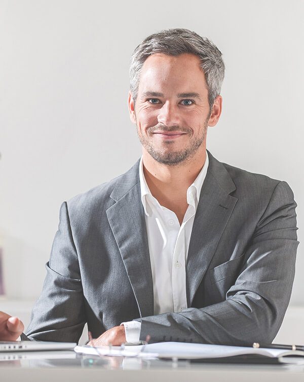 A confident man in a gray suit sitting and smiling in a bright office setting.