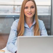 A young woman with long hair sitting at a desk with a laptop, smiling at the camera in a modern office setting.