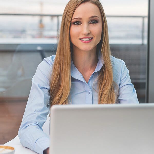 A young woman with long hair sitting at a desk with a laptop, smiling at the camera in a modern office setting.