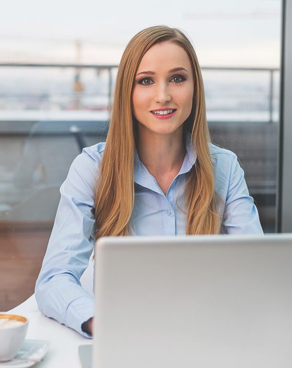 A young woman with long hair sitting at a desk with a laptop, smiling at the camera in a modern office setting.