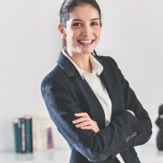 A confident businesswoman in a dark blazer stands with arms crossed, smiling in a bright office setting.