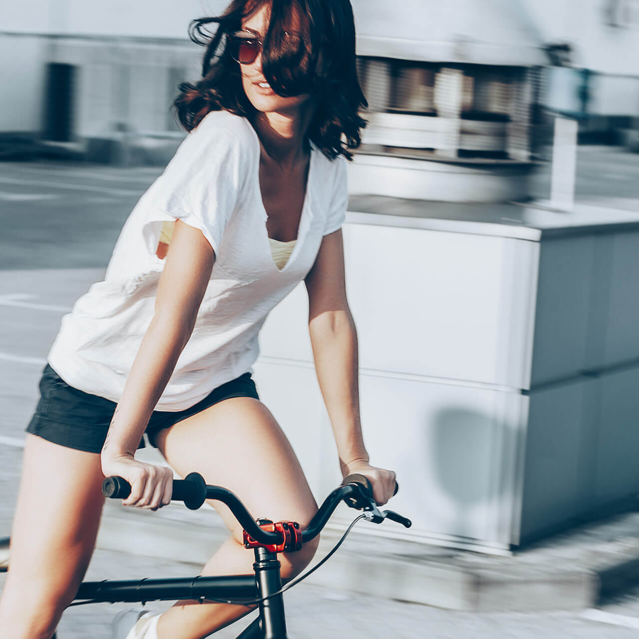 A woman with sunglasses riding a bicycle in an urban setting, wearing a white t-shirt and black shorts.